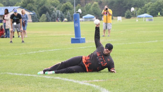 photo of Blake Boudreaux lying on his left side and holding the ball up in the air as he makes an out.  He's dressed for baseball with an eye mask.  Behind him is a large padded post that marks the nearest base.
